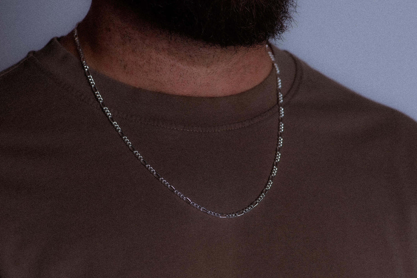 A close-up of a person wearing a silver chain necklace over a brown crewneck t-shirt, against a plain light gray background.