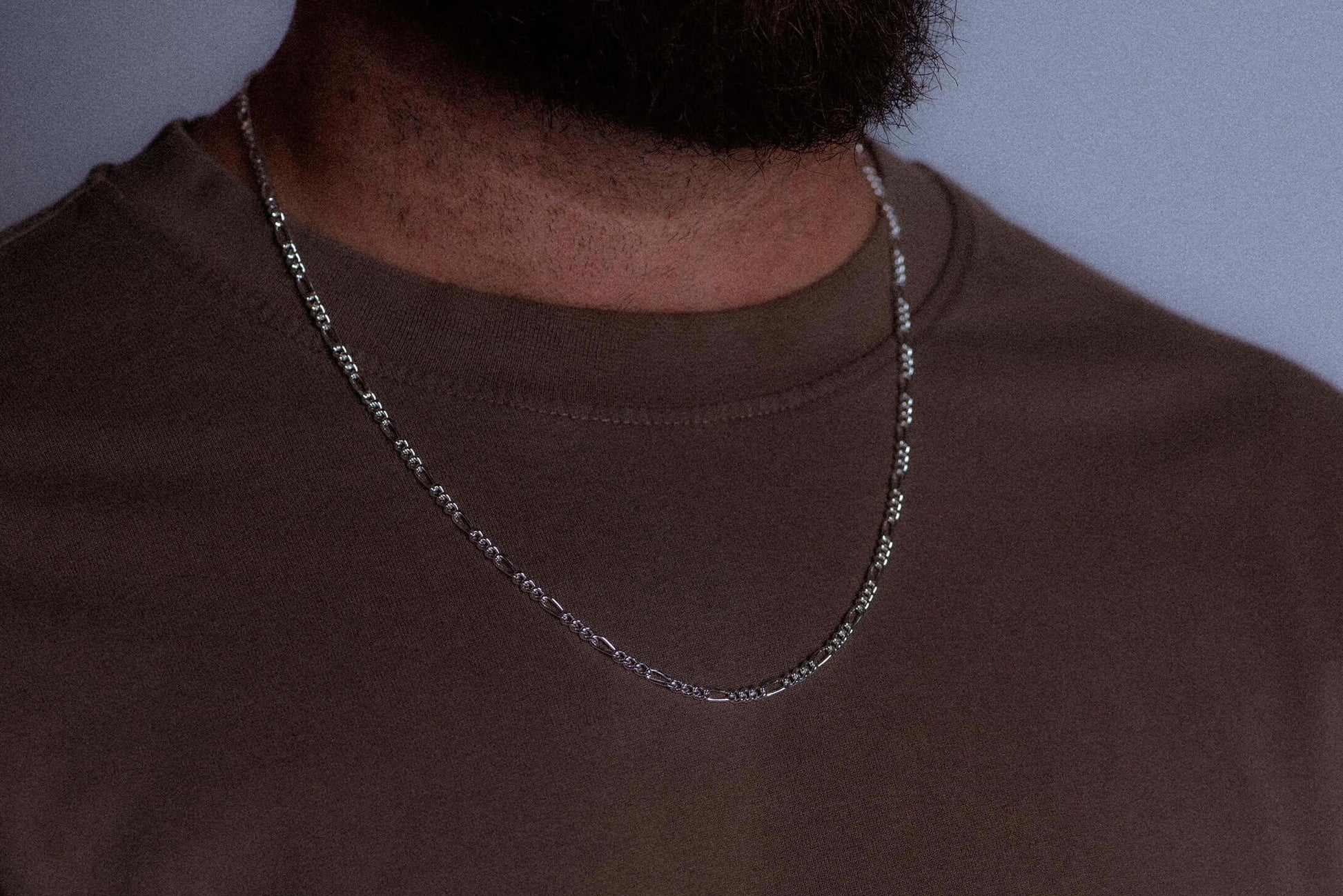 A close-up of a person wearing a silver chain necklace over a brown crewneck t-shirt, against a plain light gray background.