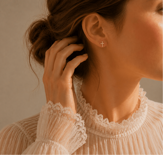 A close-up lifestyle shot of a woman wearing a delicate gold cross stud earring, touching her hair. She is wearing a cream-colored lace-trimmed blouse against a warm beige wall with soft window light shadows.